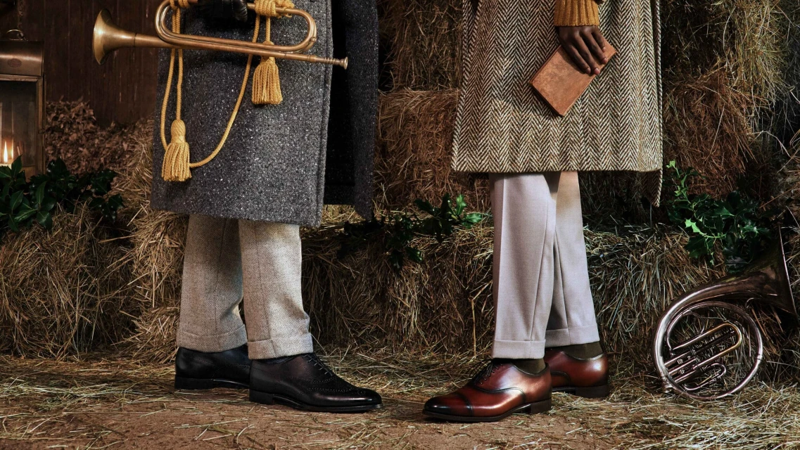 Two people stand in front of hay bales, wearing coats, trousers, and dress shoes. One holds a book, the other a trumpet.