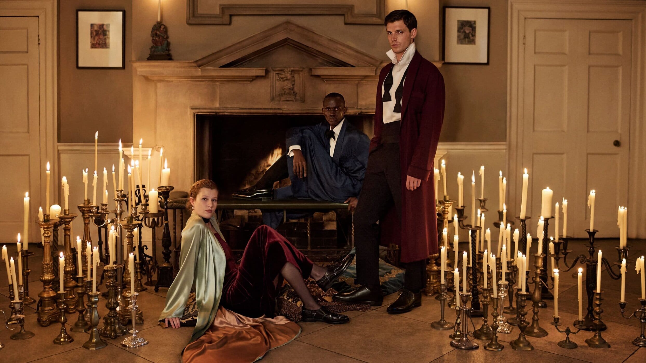 Three people are in a room filled with many lit candles on candelabras. Two men in suits and a woman in dress attire. Fireplace in background.