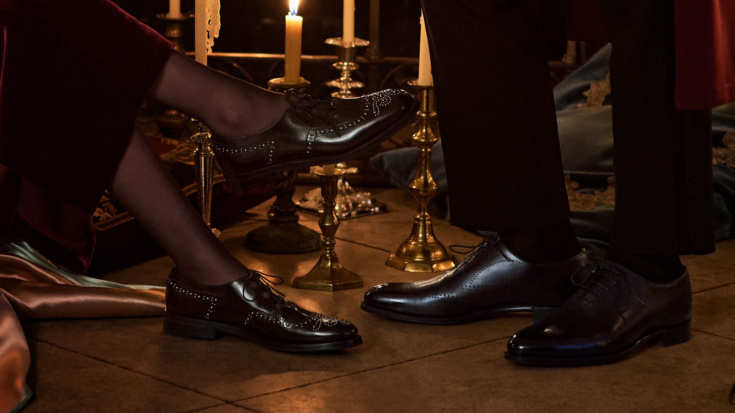 Eye-level shot of a woman's and man's legs and black dress shoes, set on a tile floor next to brass candle holders and lit candles.
