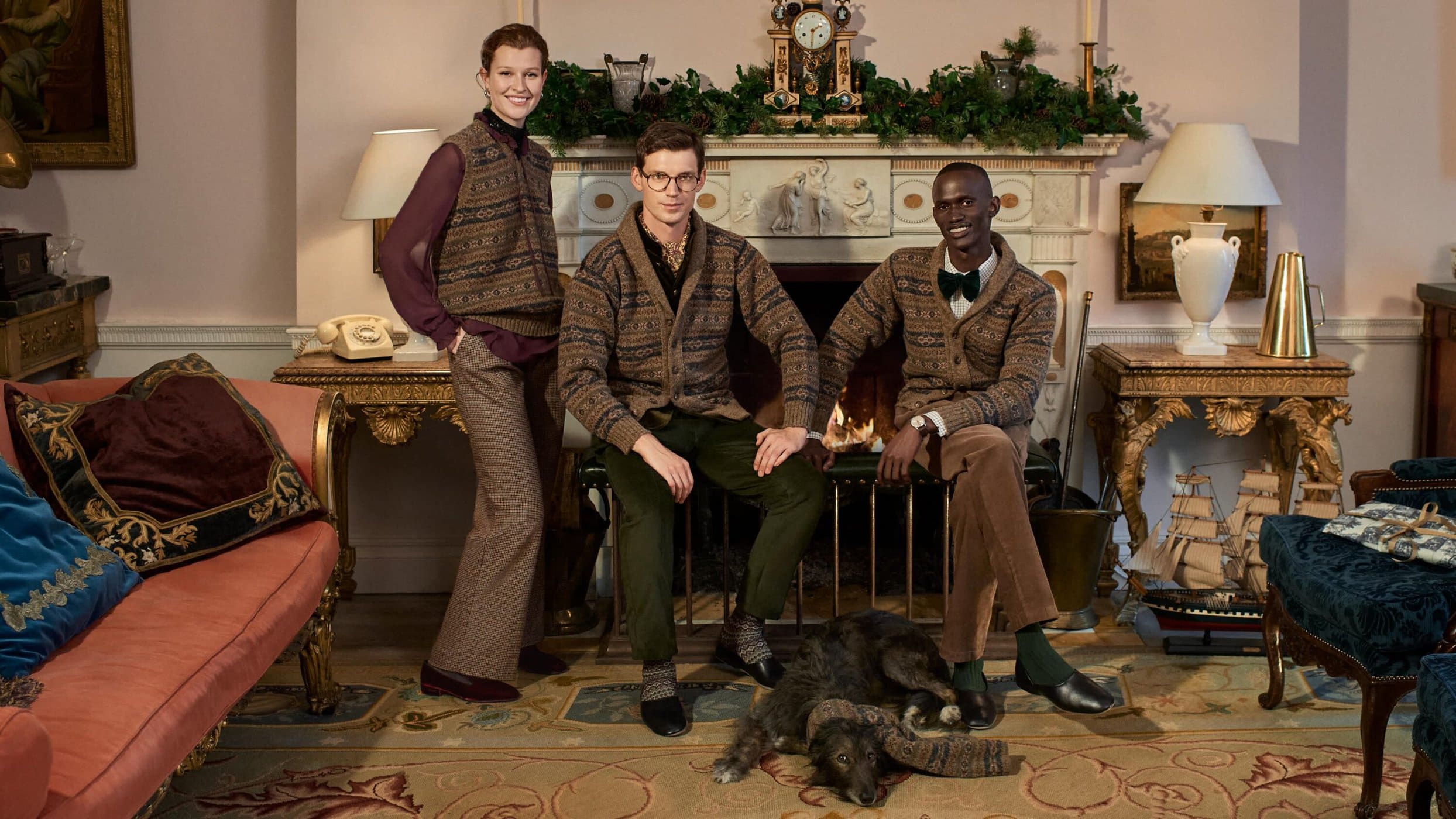 Three people in patterned knitwear pose indoors with a dog near a fireplace and furniture; fashion, lifestyle, interior design.