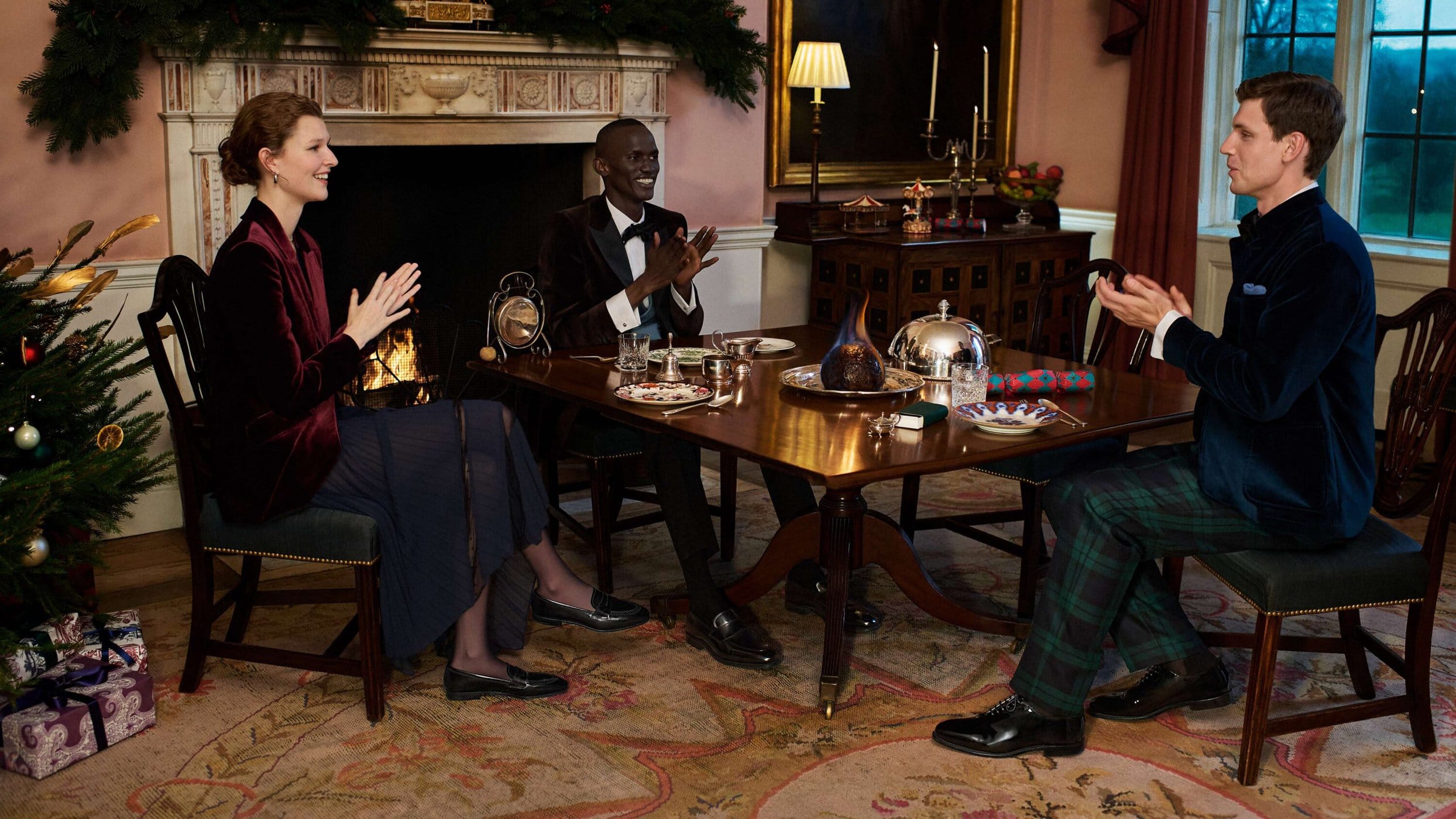 Three people in formal attire are seated at a wooden table with place settings. A Christmas tree and fireplace add to the festive scene.