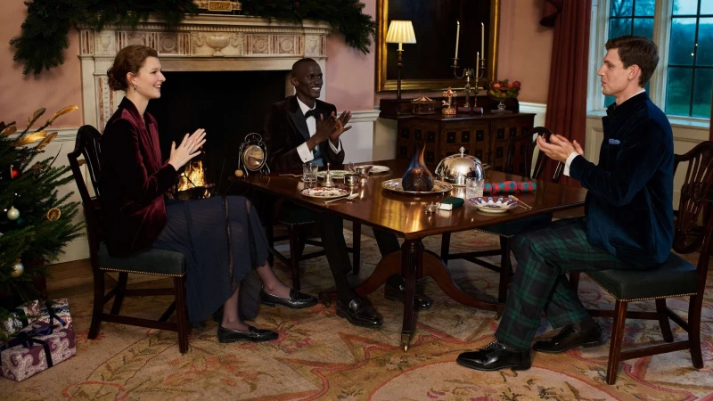 Three people in formal attire are seated at a wooden table with place settings. A Christmas tree and fireplace add to the festive scene.