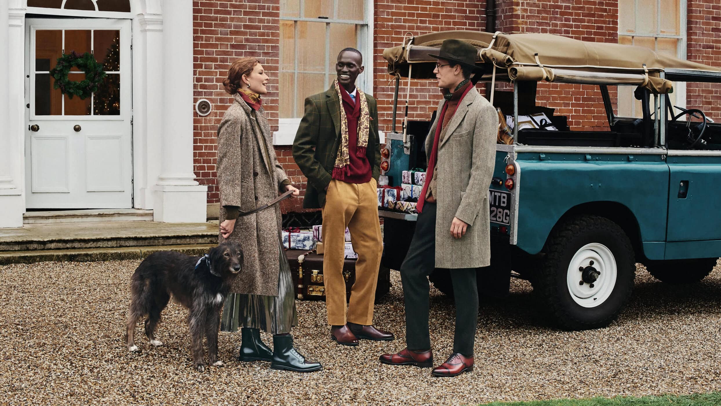 Three people and a dog pose in front of a vintage SUV, stone building, and decorated door for a fashion lifestyle editorial.