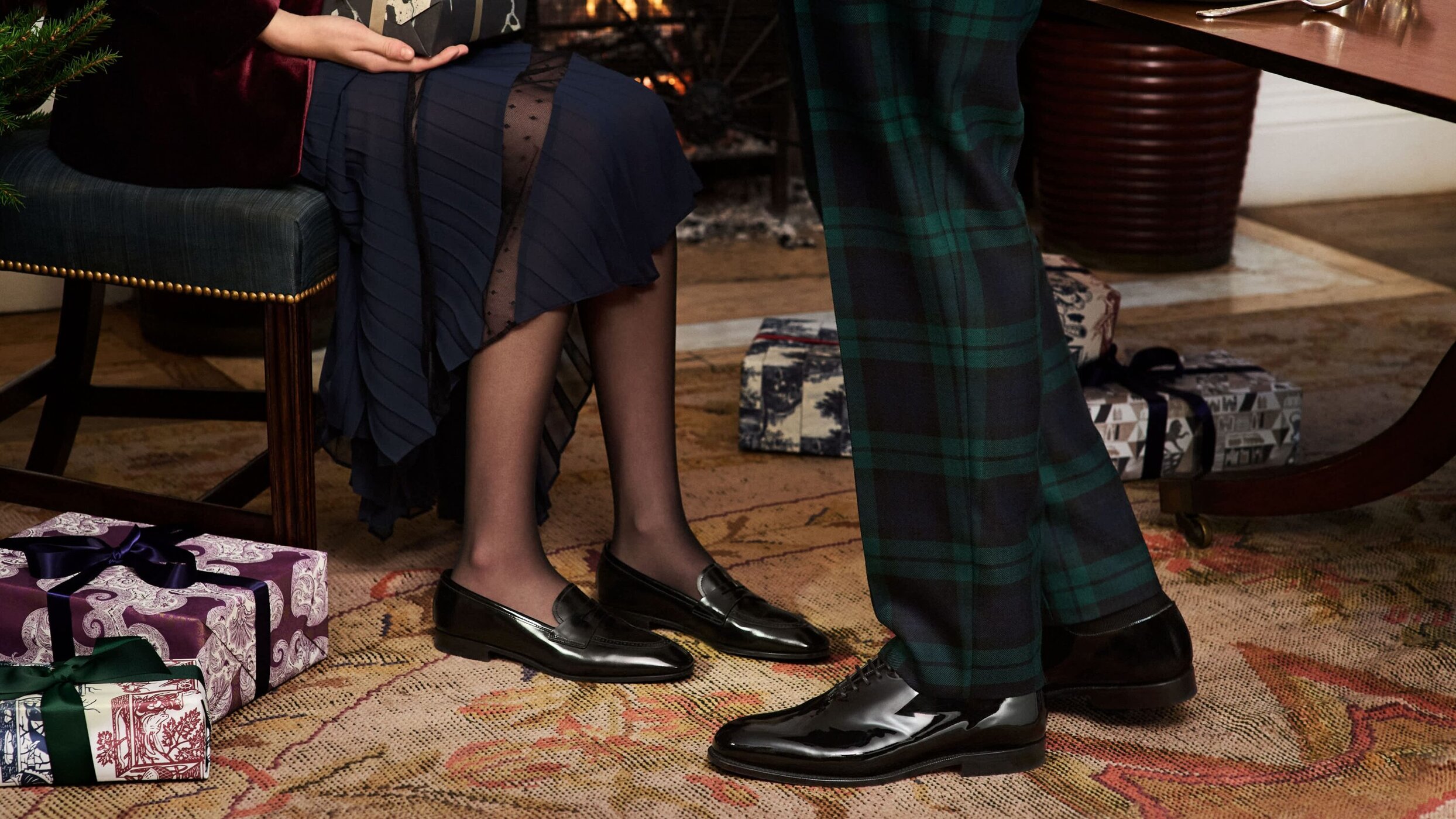 A couple is indoors for the holidays near a fireplace, surrounded by beautifully wrapped presents and wearing stylish shoes and socks.