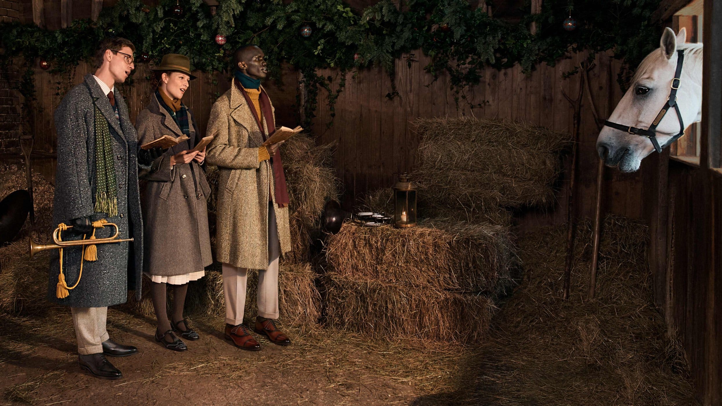 Three people in vintage clothing hold books and a trumpet in a stable next to a white horse, with garland and decorations hanging.