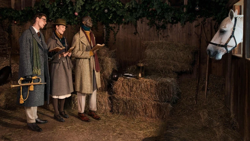 Three people in vintage clothing hold books and a trumpet in a stable next to a white horse, with garland and decorations hanging.