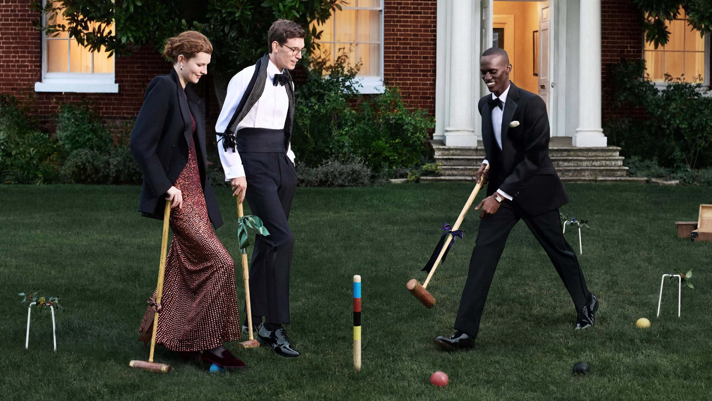 Three people in formal wear playing croquet on a lawn in front of a brick building, illuminated by natural light.