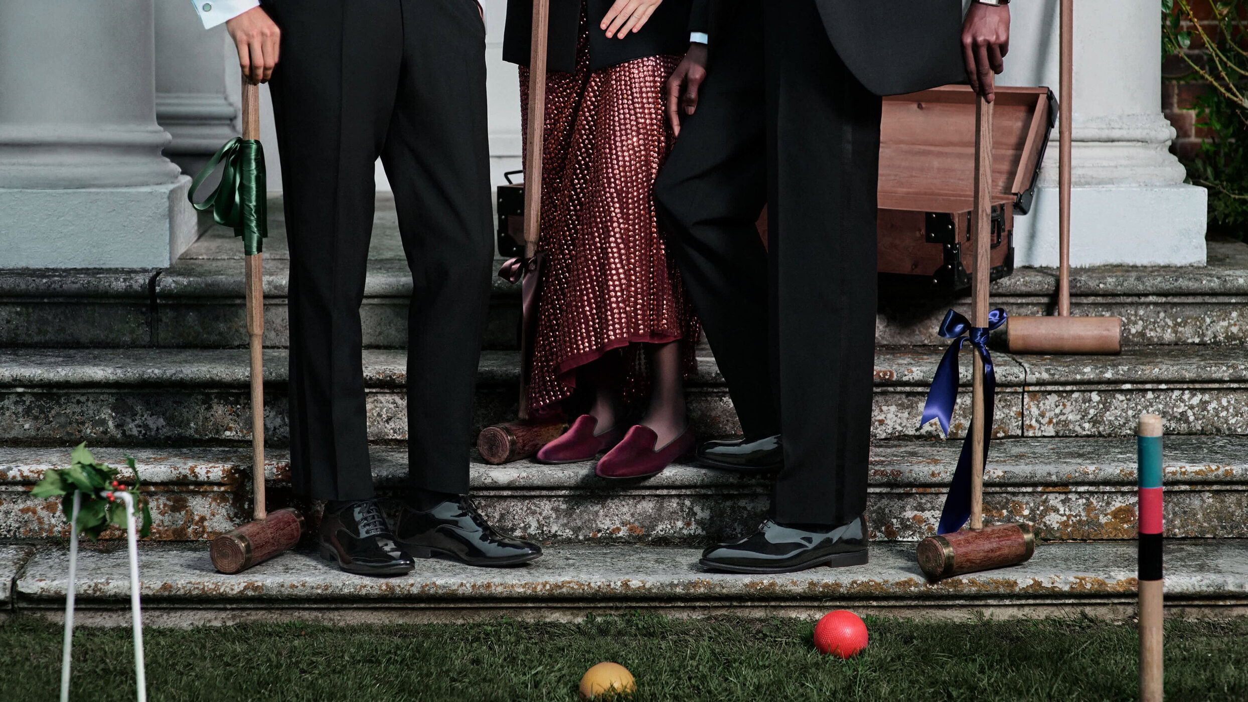 Croquet game on lawn: three people in formal attire stand on stone steps holding mallets; vintage trunk, lawn, green grass, brick building.