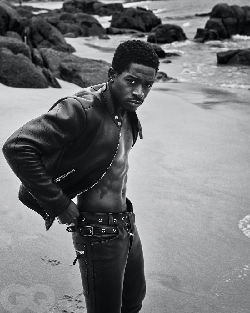 Monochrome portrait of a Black male model with a serious expression, wearing a leather jacket and pants on a sandy beach with rocky coastline.