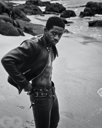 Monochrome portrait of a Black male model with a serious expression, wearing a leather jacket and pants on a sandy beach with rocky coastline.