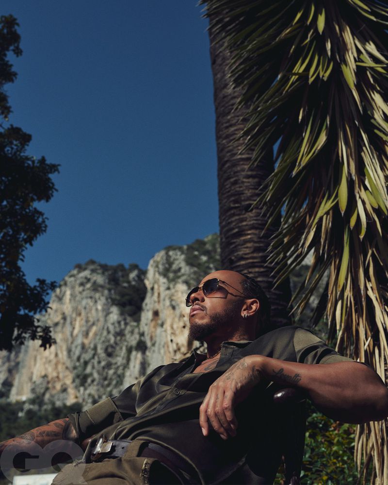 Lewis Hamilton poses in an olive green shirt and sunglasses in a portrait featuring a palm tree and mountain background in daylight.