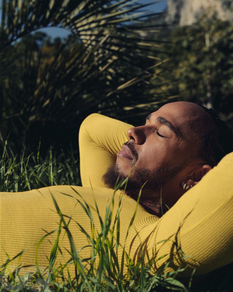 Adult male with eyes closed reclines in grass wearing yellow top, sunlight on his face, trees and foliage in soft focus background.
