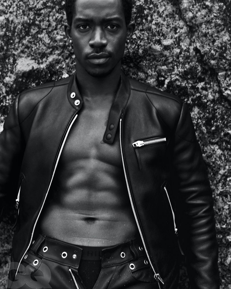 Black and white portrait of a muscular Black man in a leather jacket and leather pants, standing in front of a rough rock surface.