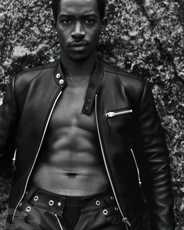 Black and white portrait of a muscular Black man in a leather jacket and leather pants, standing in front of a rough rock surface.