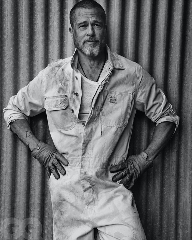 Black and white portrait of Brad Pitt wearing a light-colored jumpsuit and gloves, standing against a corrugated metal wall background.