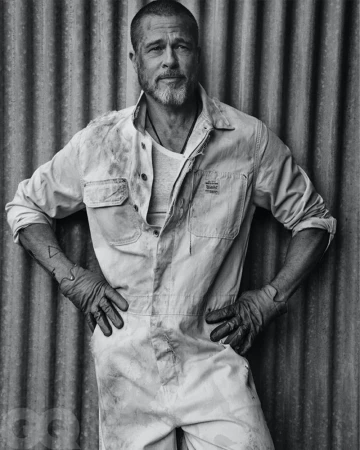 Black and white portrait of Brad Pitt wearing a light-colored jumpsuit and gloves, standing against a corrugated metal wall background.