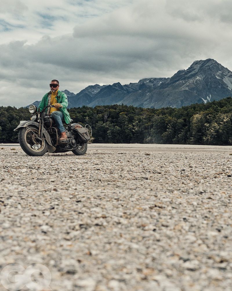 Adult male riding vintage motorcycle on a gravel plain with mountains and cloudy sky in the background in an eye-level medium shot.