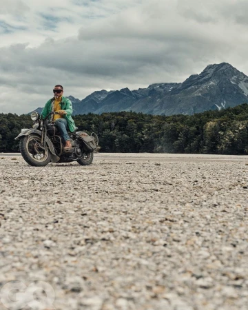 Adult male riding vintage motorcycle on a gravel plain with mountains and cloudy sky in the background in an eye-level medium shot.