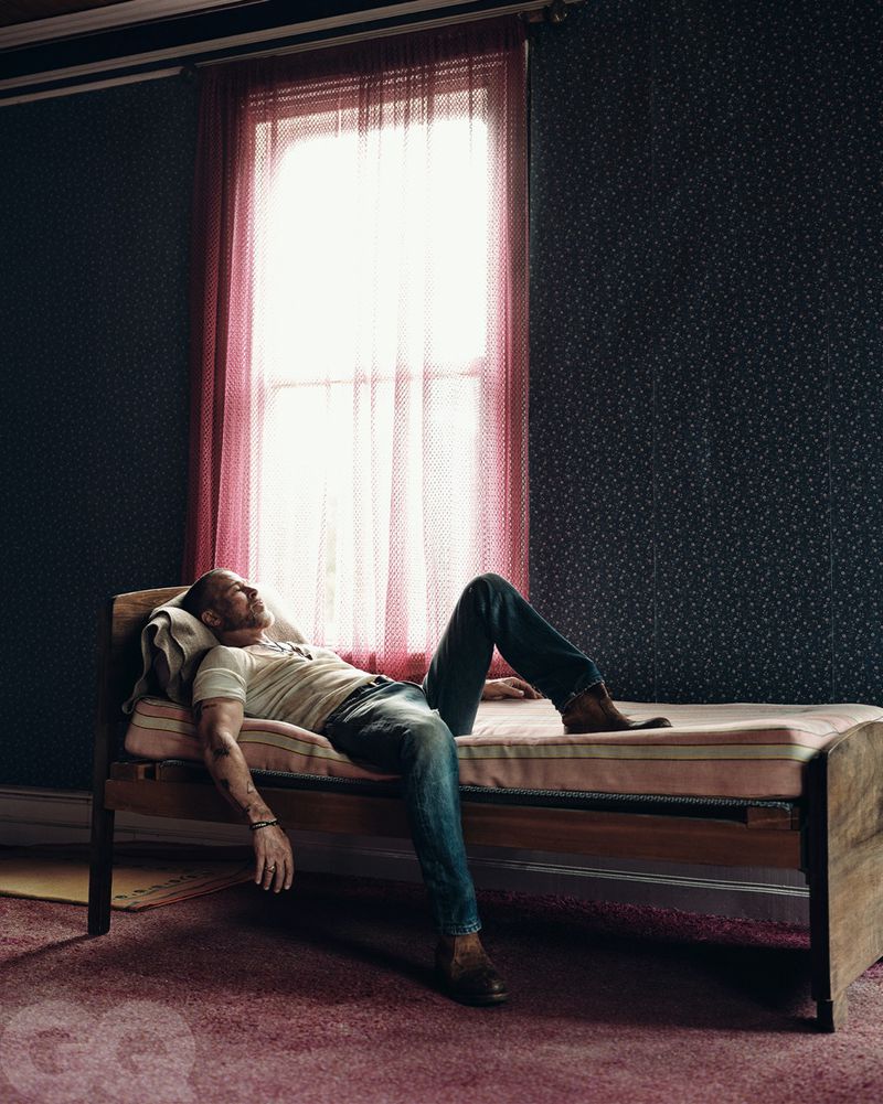 Man lounges on a daybed in a room with patterned wallpaper, illuminated by window light with sheer curtains, in a casual editorial style.