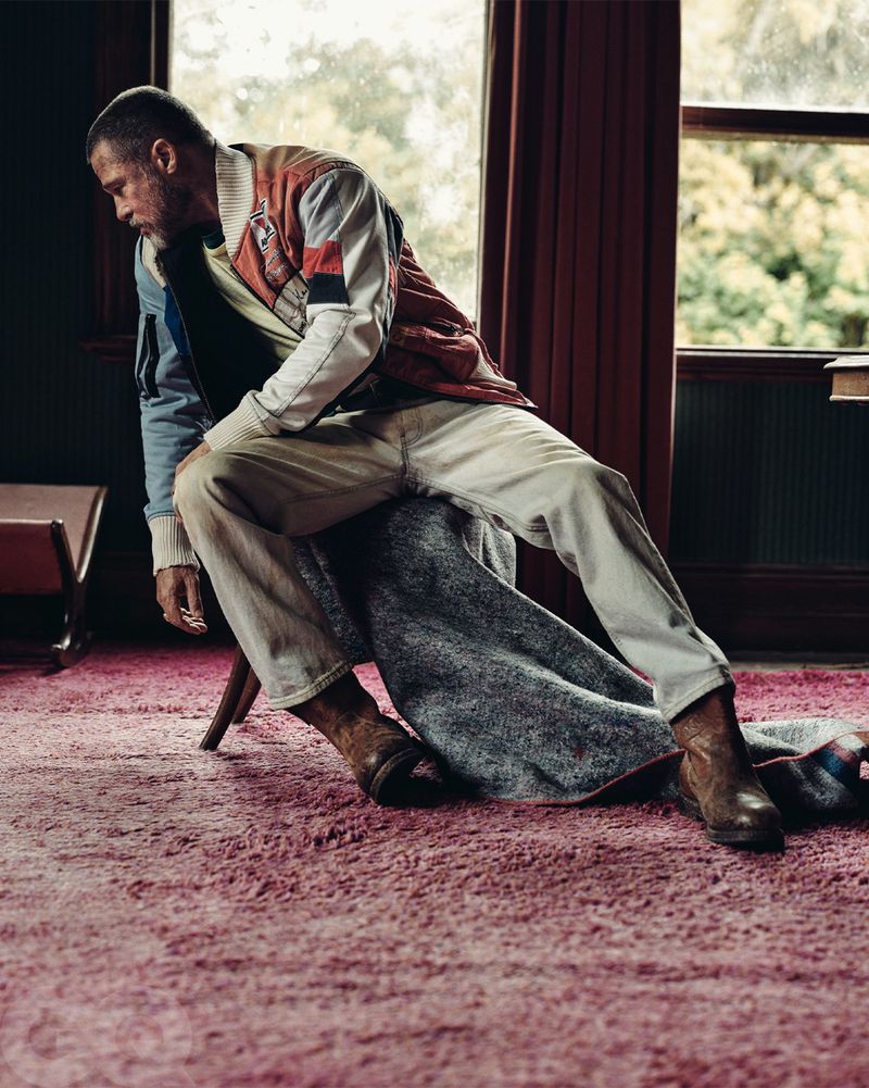 Adult male model sitting on a stool in a colorful jacket, jeans, and brown cowboy boots indoors with a window and red drapes.
