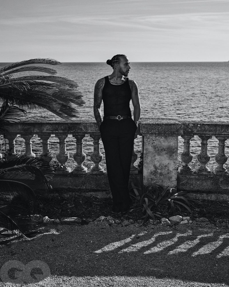 Black and white portrait of a man with a bun standing on a terrace overlooking the ocean with palm trees on the left.