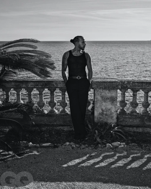 Black and white portrait of a man with a bun standing on a terrace overlooking the ocean with palm trees on the left.