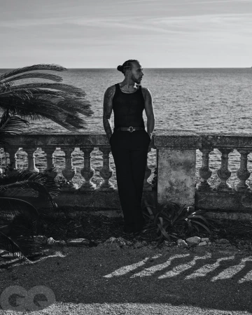 Black and white portrait of a man with a bun standing on a terrace overlooking the ocean with palm trees on the left.