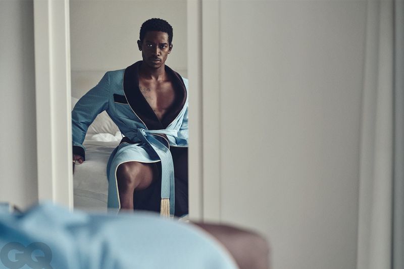 Adult male poses in a light blue robe, half of his body shown in a reflection of a mirror inside a bedroom setting.