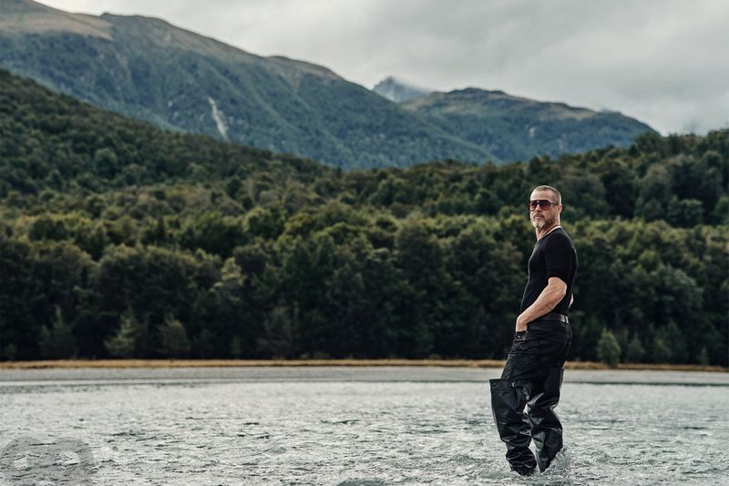 Adult male standing waist-deep in water with mountains and forest in the background on a cloudy day, wearing sunglasses and waders.