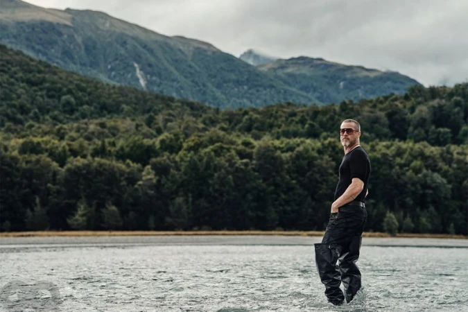 Adult male standing waist-deep in water with mountains and forest in the background on a cloudy day, wearing sunglasses and waders.