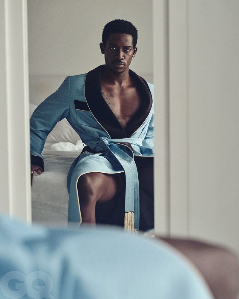Portrait of a Black man in a light blue robe sitting on a bed, seen through a mirror in an indoor setting with natural lighting.