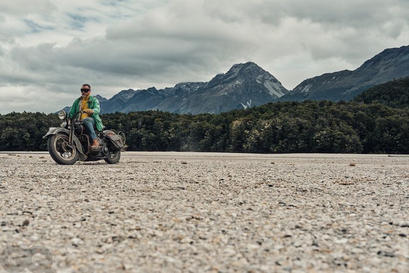 Man on a vintage motorcycle in a vast, rocky landscape with mountains in the background under a cloudy sky, creating a sense of adventure.