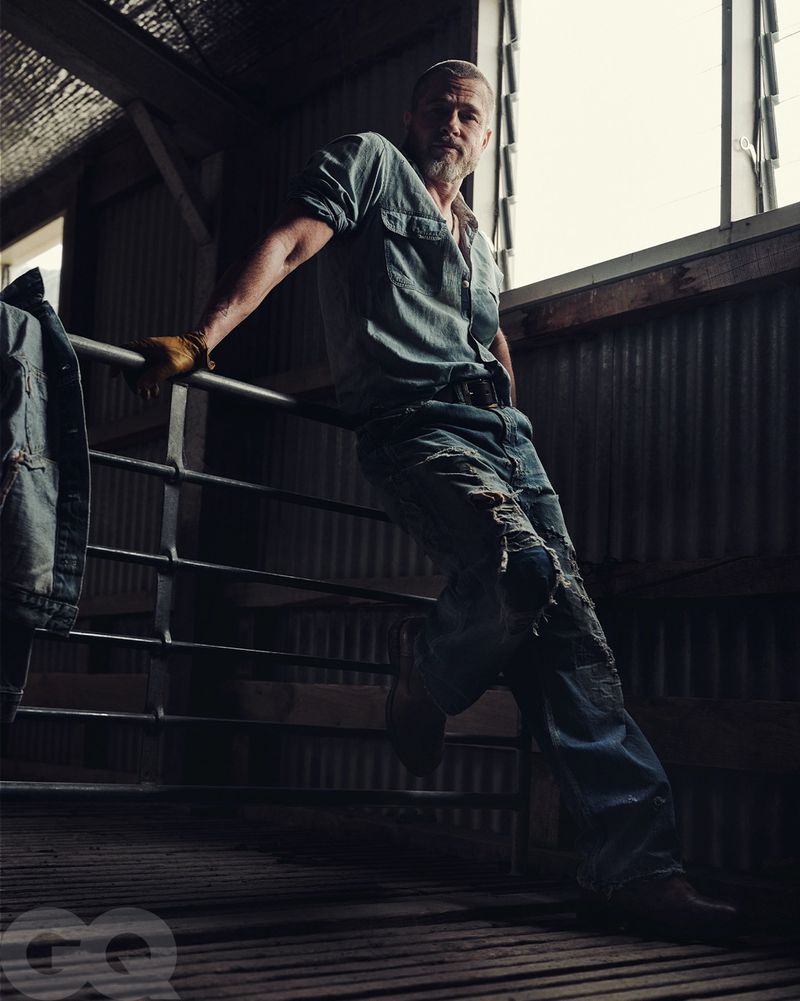 Portrait of adult male in denim shirt and jeans, leaning on a metal railing indoors with natural window light, with a 'GQ' logo.