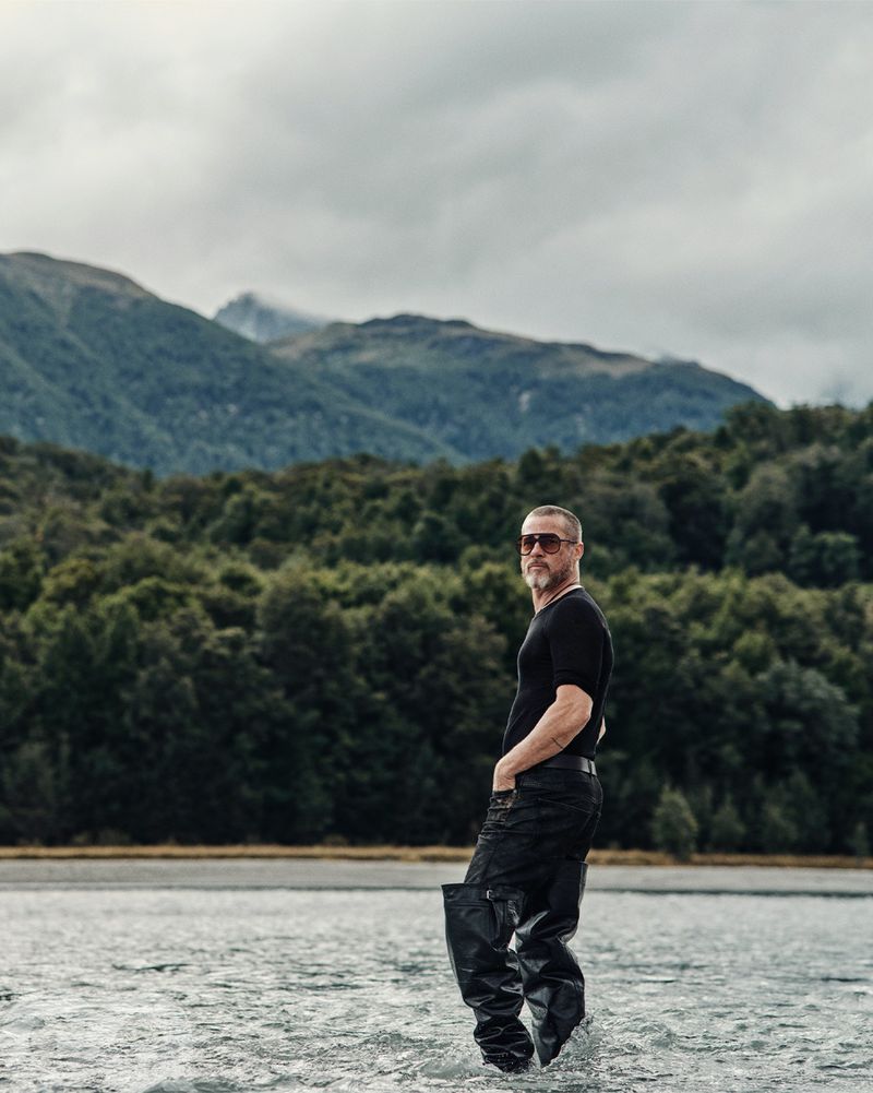 Man in waders standing in a lake, mountains and forest in the background, overcast sky, full shot, outdoor setting.
