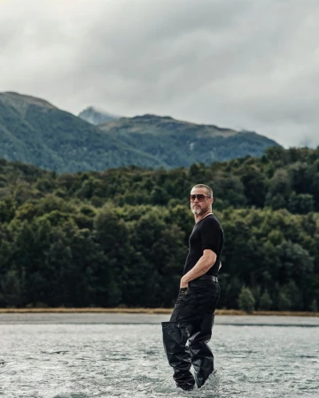 Man in waders standing in a lake, mountains and forest in the background, overcast sky, full shot, outdoor setting.