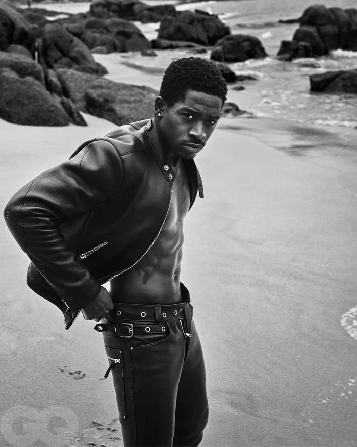 Damson Idris wearing a leather jacket and trousers standing on a rocky beach in a black and white editorial photograph.