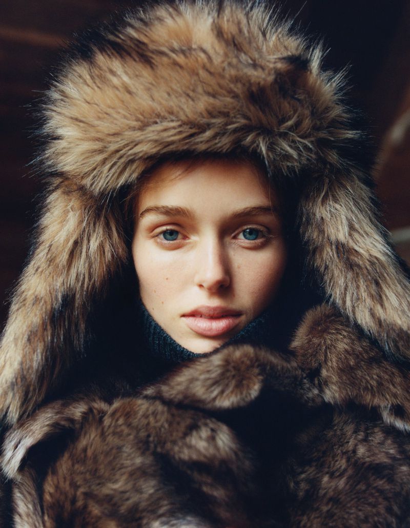 Close-up portrait of a woman with blue eyes wearing a fur hat and turtleneck sweater, captured in soft, warm lighting.