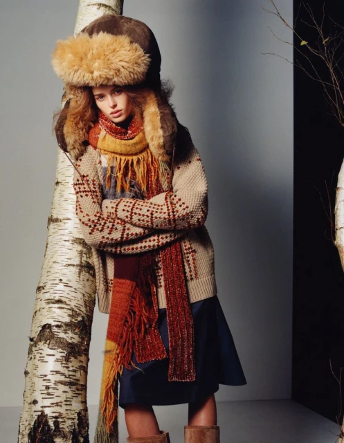 A woman in a fur hat, patterned sweater and scarf leans against a birch tree, posing in a fashion editorial with a grey background.