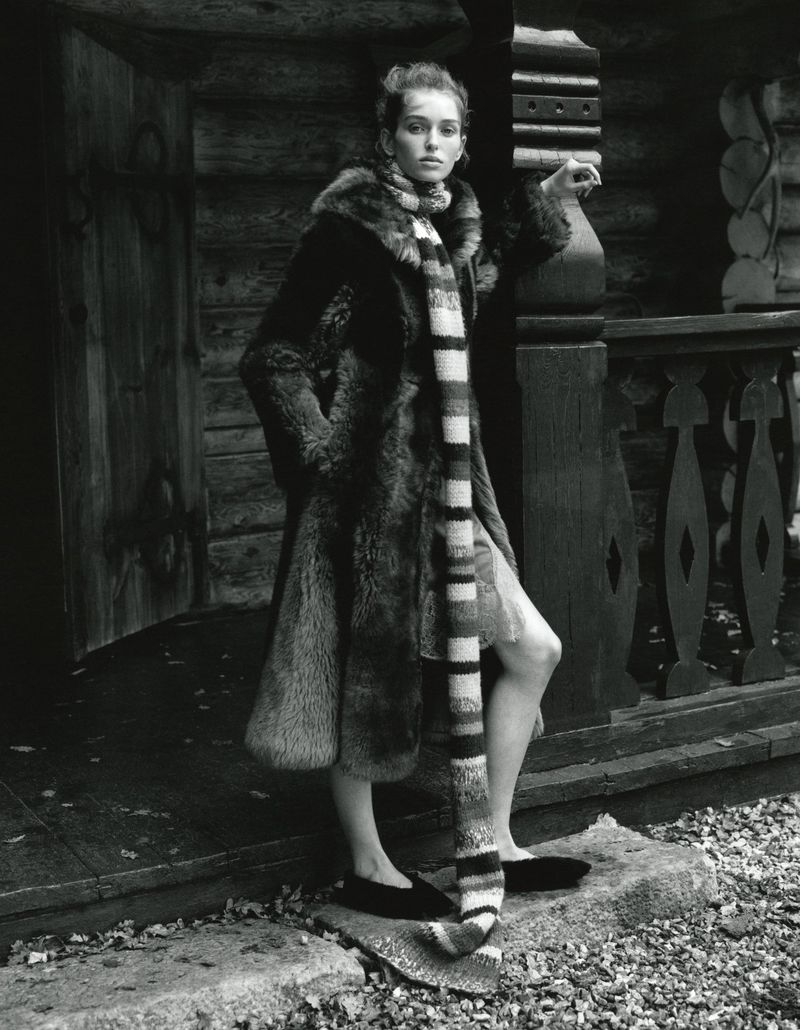 Monochrome portrait of a woman in a fur coat and striped scarf standing outside a log cabin, fashion editorial style.