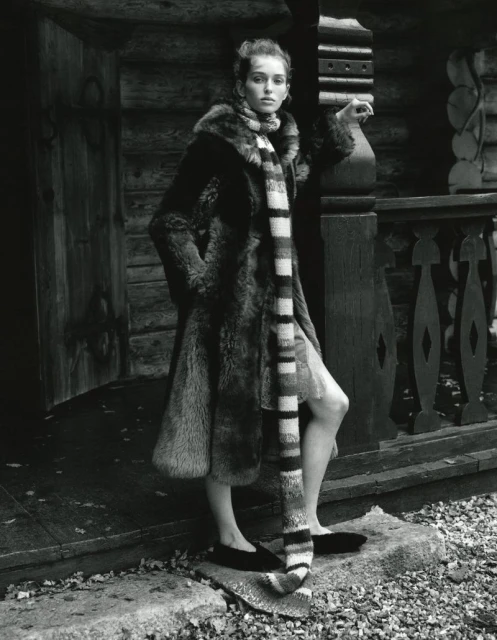 Monochrome portrait of a woman in a fur coat and striped scarf standing outside a log cabin, fashion editorial style.
