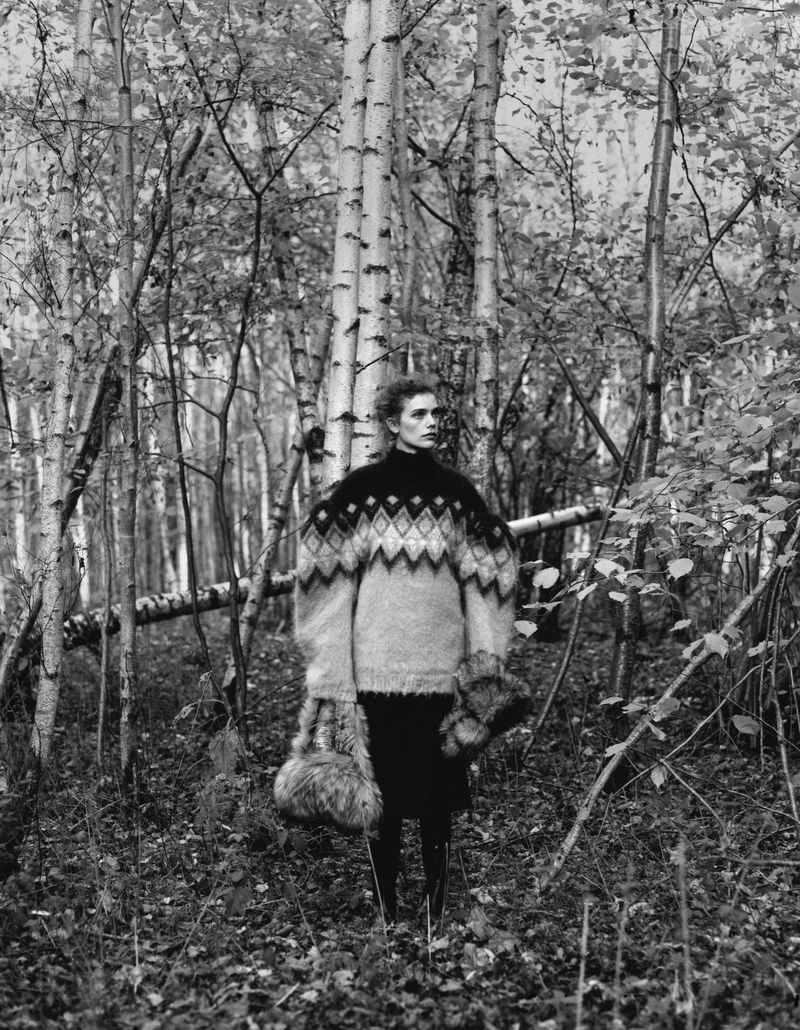 Monochrome fashion shot of woman in oversized patterned sweater in forest setting with birch trees, holding fur accessories, editorial style.