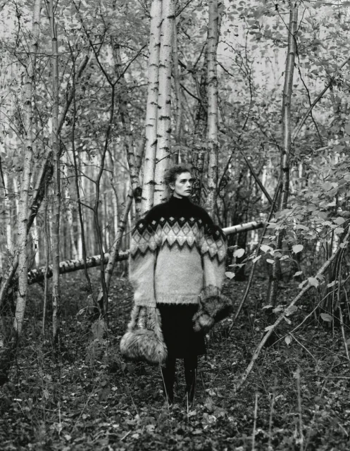 Monochrome fashion shot of woman in oversized patterned sweater in forest setting with birch trees, holding fur accessories, editorial style.