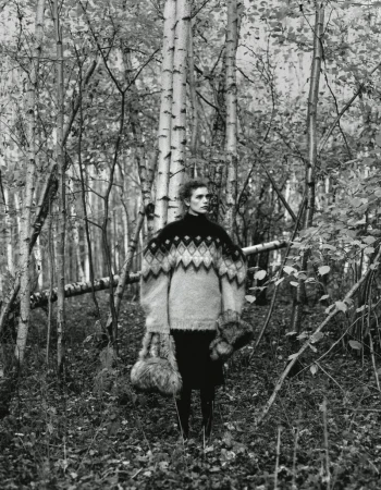Monochrome fashion shot of woman in oversized patterned sweater in forest setting with birch trees, holding fur accessories, editorial style.