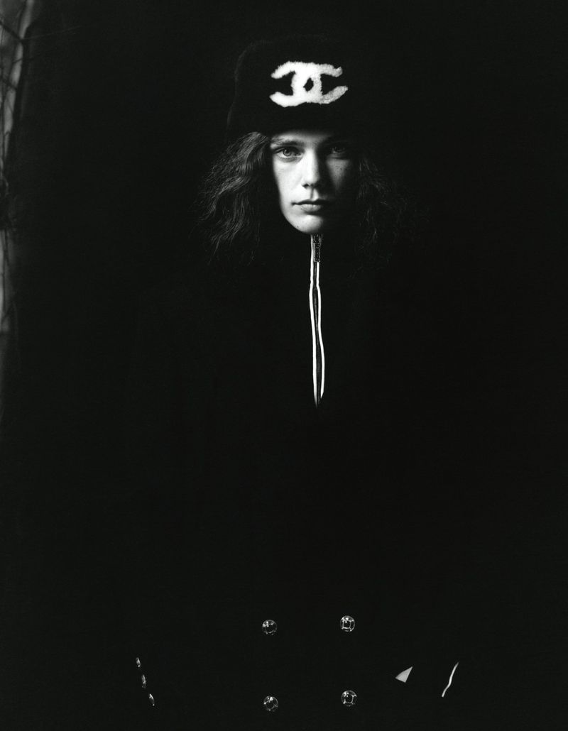 A portrait-style monochrome photograph features a young adult wearing a Chanel logo hat and dark coat in a dimly lit setting.
