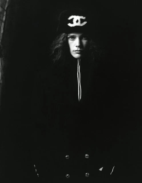 A portrait-style monochrome photograph features a young adult wearing a Chanel logo hat and dark coat in a dimly lit setting.
