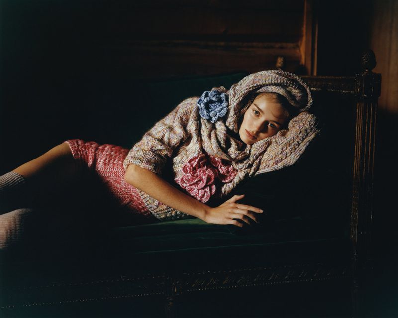 A fair-skinned adult female model with wavy blonde hair lies on a velvet couch wearing a knit sweater and skirt with floral decorations.