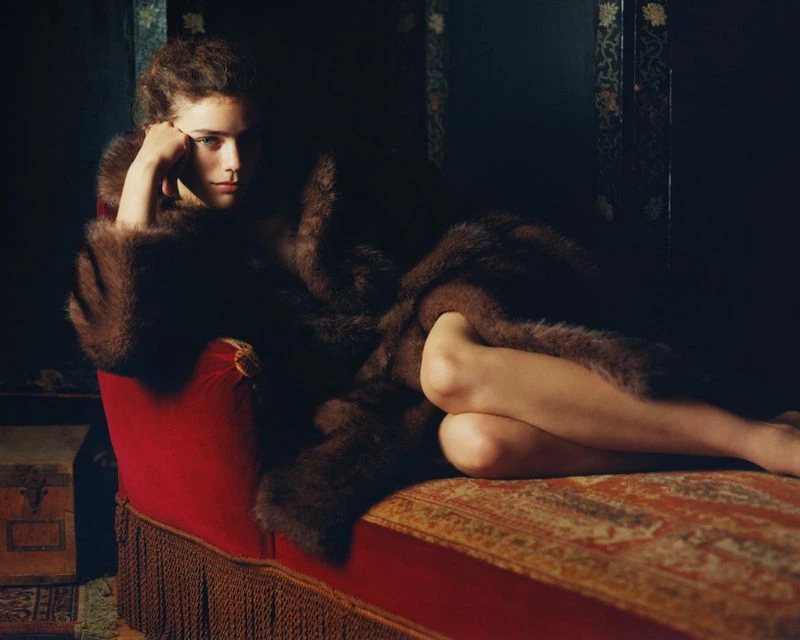 Woman with curly hair reclines on a red chaise lounge wearing a dark brown fur coat in a dimly lit indoor setting.