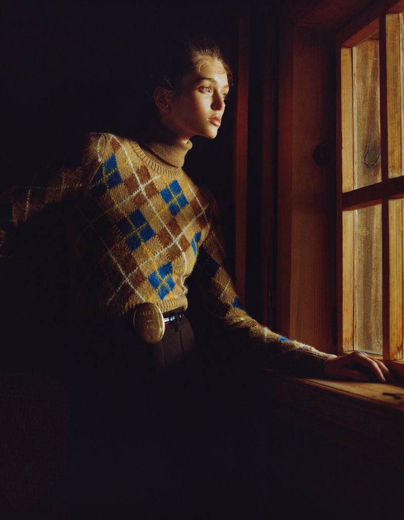 Woman in argyle sweater looking out of a window in a dark room, illuminated by a warm light with high contrast and shallow depth of field.