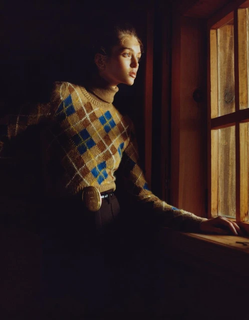 Woman in argyle sweater looking out of a window in a dark room, illuminated by a warm light with high contrast and shallow depth of field.