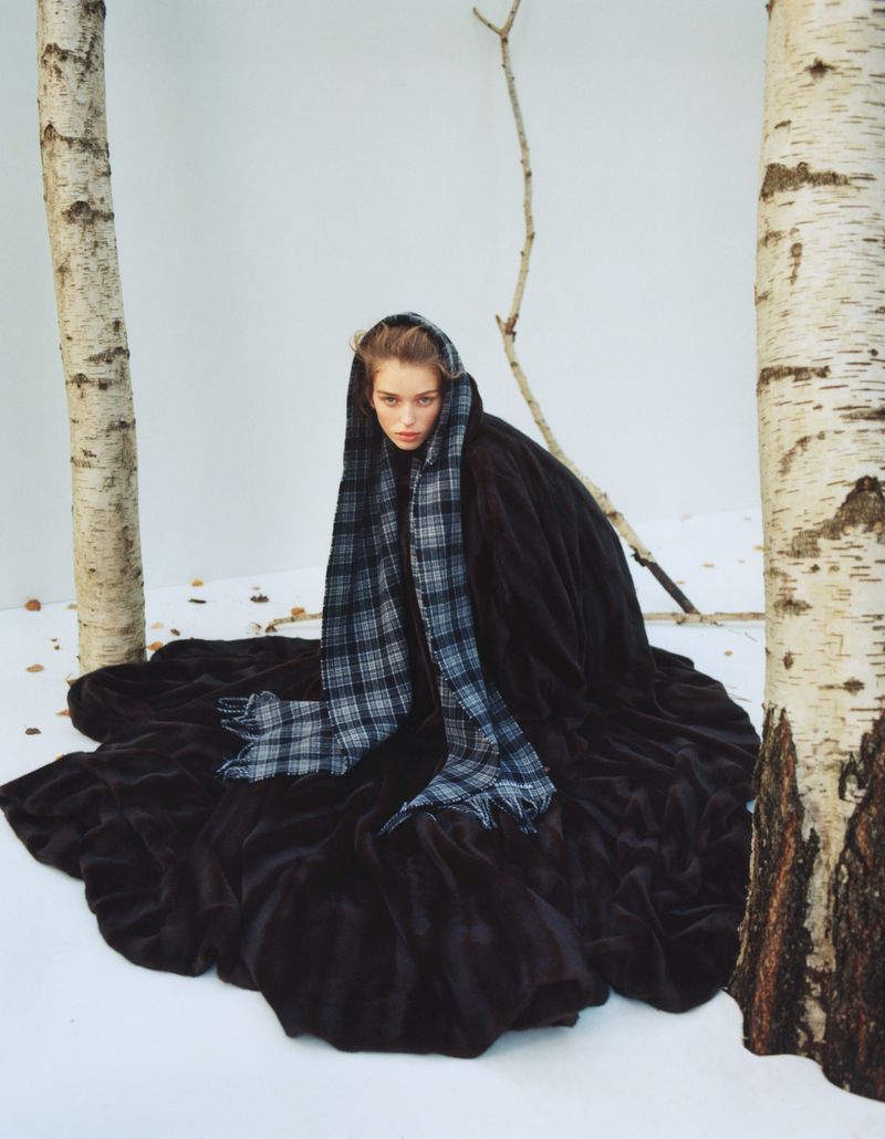A woman draped in dark fabric and a plaid scarf sits among birch trees, a striking fashion image in an outdoor setting.